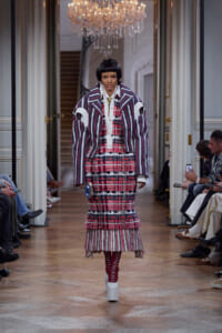 Model walks a runway in a bold layered plaid outfit with a striped oversized jacket, white shirt, and red–pink plaid skirt in a chandelier-lit hall.