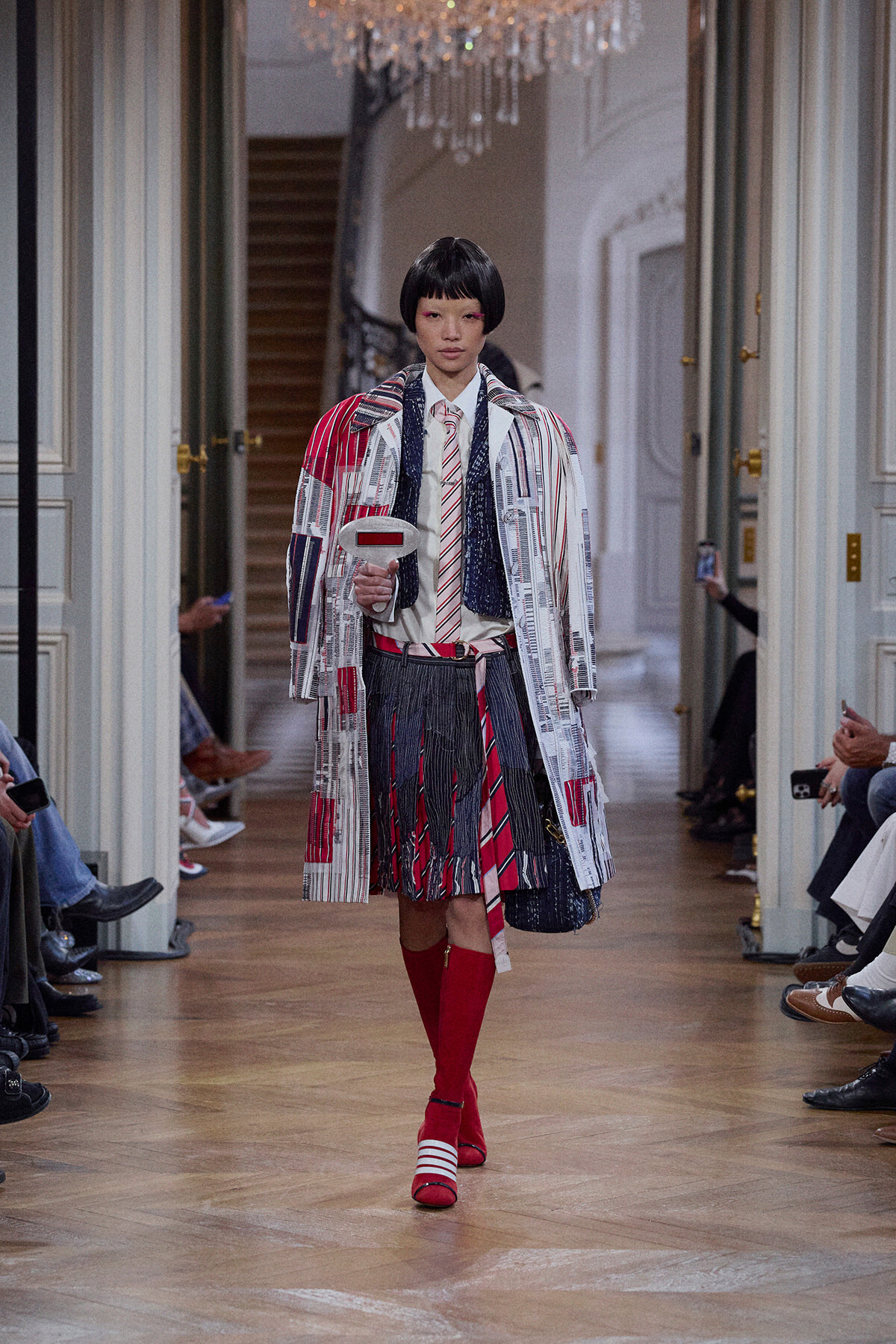 Model on a runway wearing a bold patchwork coat, white shirt with a striped tie, and bright red knee-high socks.