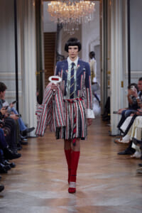 Model walking a fashion runway in a blue striped blazer, matching striped skirt, and red knee-high socks; holds a white oval sign and draped striped coat, chandeliers overhead.
