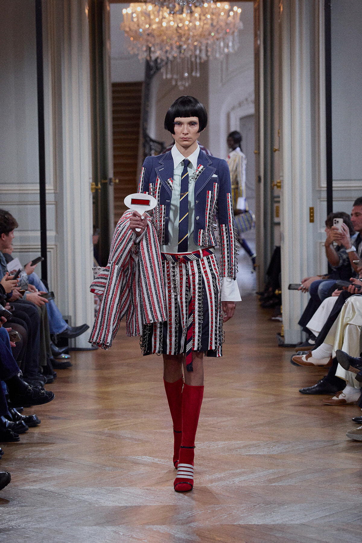 Model walking a fashion runway in a blue striped blazer, matching striped skirt, and red knee-high socks; holds a white oval sign and draped striped coat, chandeliers overhead.