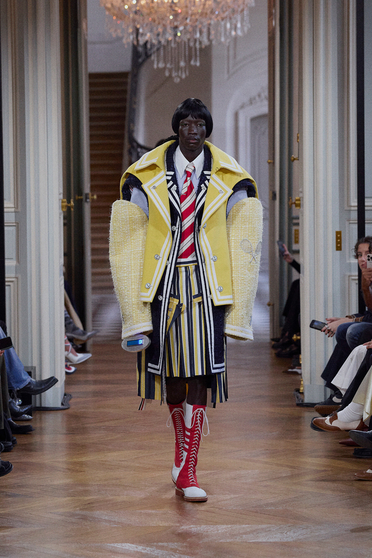 Model on a runway wearing a bright yellow oversized coat with white trim over a striped suit, paired with red knee-high lace-up boots.