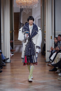 Model walking a runway in an oversized white coat with navy trim, layered blue knitwear, and lime knee-high boots, holding a blue device in front.
