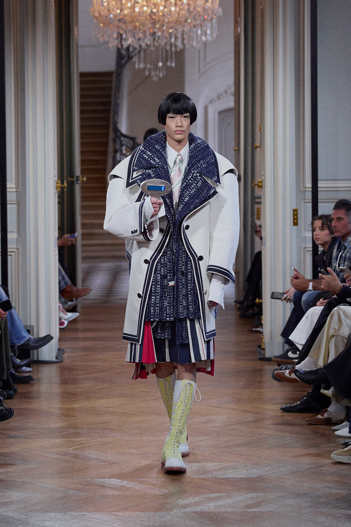 Model walking a runway in an oversized white coat with navy trim, layered blue knitwear, and lime knee-high boots, holding a blue device in front.
