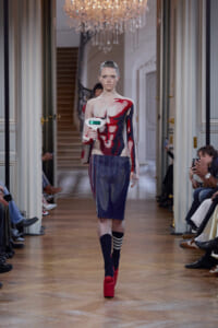 Model walks down a fashion-show runway in a graphic knit top and blue pencil skirt with red platform heels, holding a small white prop displaying a green digital readout.