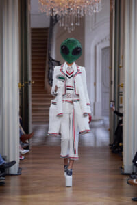 Model walks a fashion runway in a white suit with red/blue trim, wearing a glittering green alien head mask and platform heels, hallway backdrop.