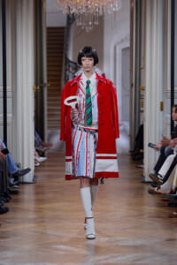 Model on a fashion runway wearing a red glossy trench over a striped suit with a green tie, holding a white oval sign in front of an audience.