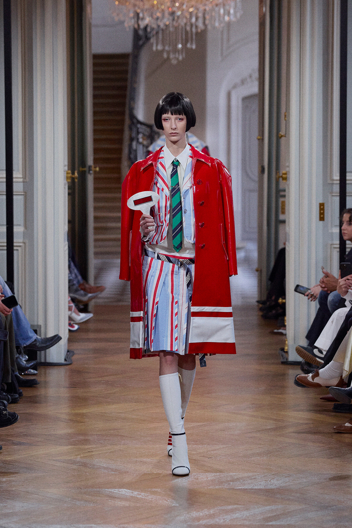 Model on a fashion runway wearing a red glossy trench over a striped suit with a green tie, holding a white oval sign in front of an audience.