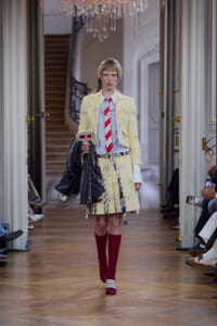 Model walks a fashion runway in a pale yellow tweed suit with a red striped tie, carrying a dark jacket and a white shirt. She wears maroon knee-high socks and white sandals within a grand, chandeliered hall.