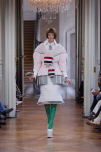 Model on a runway wearing a pale pink oversized coat with a chunky scarf, white knit skirt, and bright green lace-up boots.