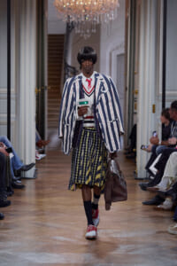 Model walks the runway in a navy-and-white striped oversized blazer, red tie, and a bold patterned midi skirt, carrying a brown bag.