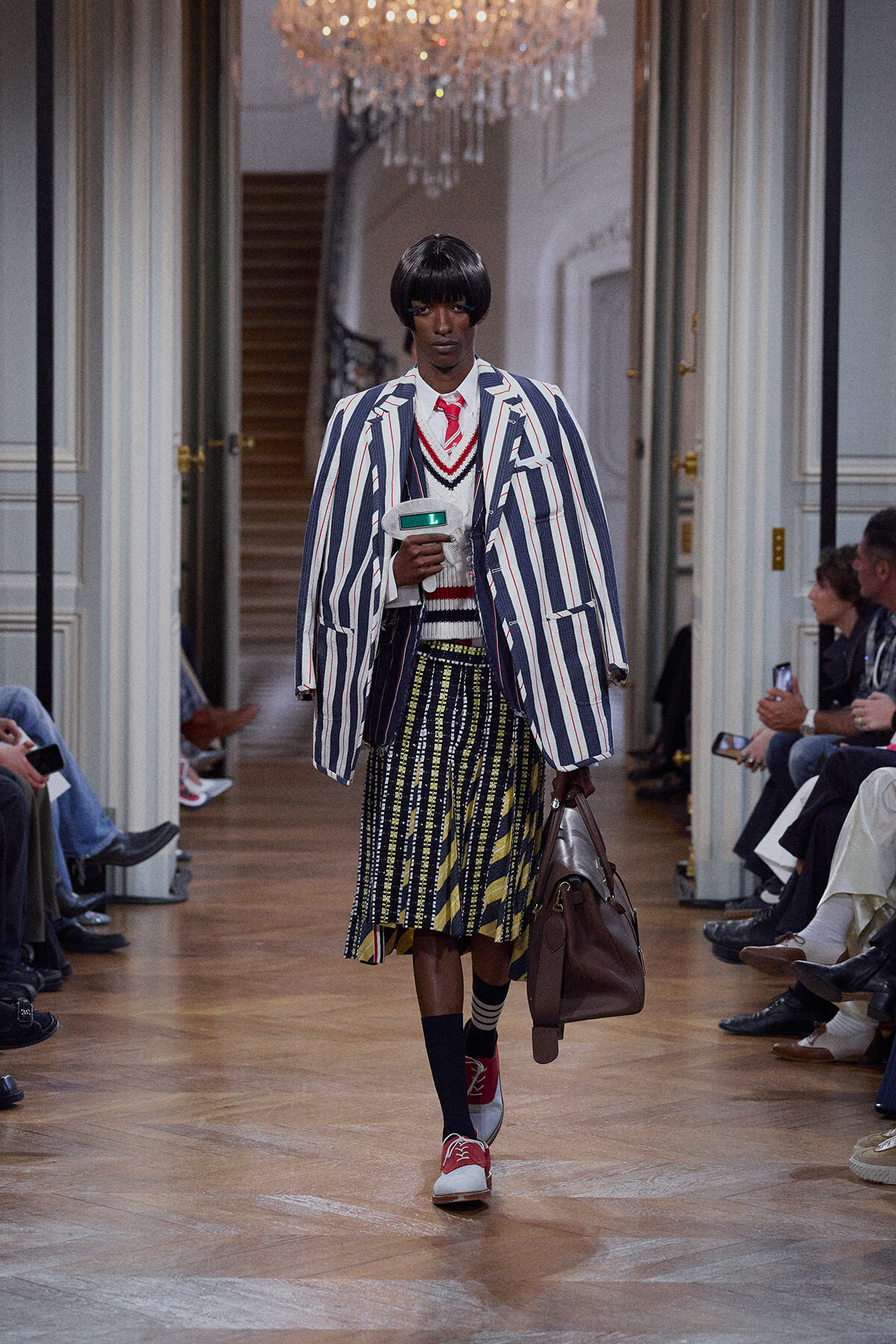Model walks the runway in a navy-and-white striped oversized blazer, red tie, and a bold patterned midi skirt, carrying a brown bag.