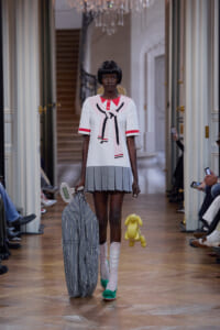 Model on a runway wearing a white top with a bow design and a pleated gray mini-skirt, carrying a large striped bag and a yellow plush bunny.