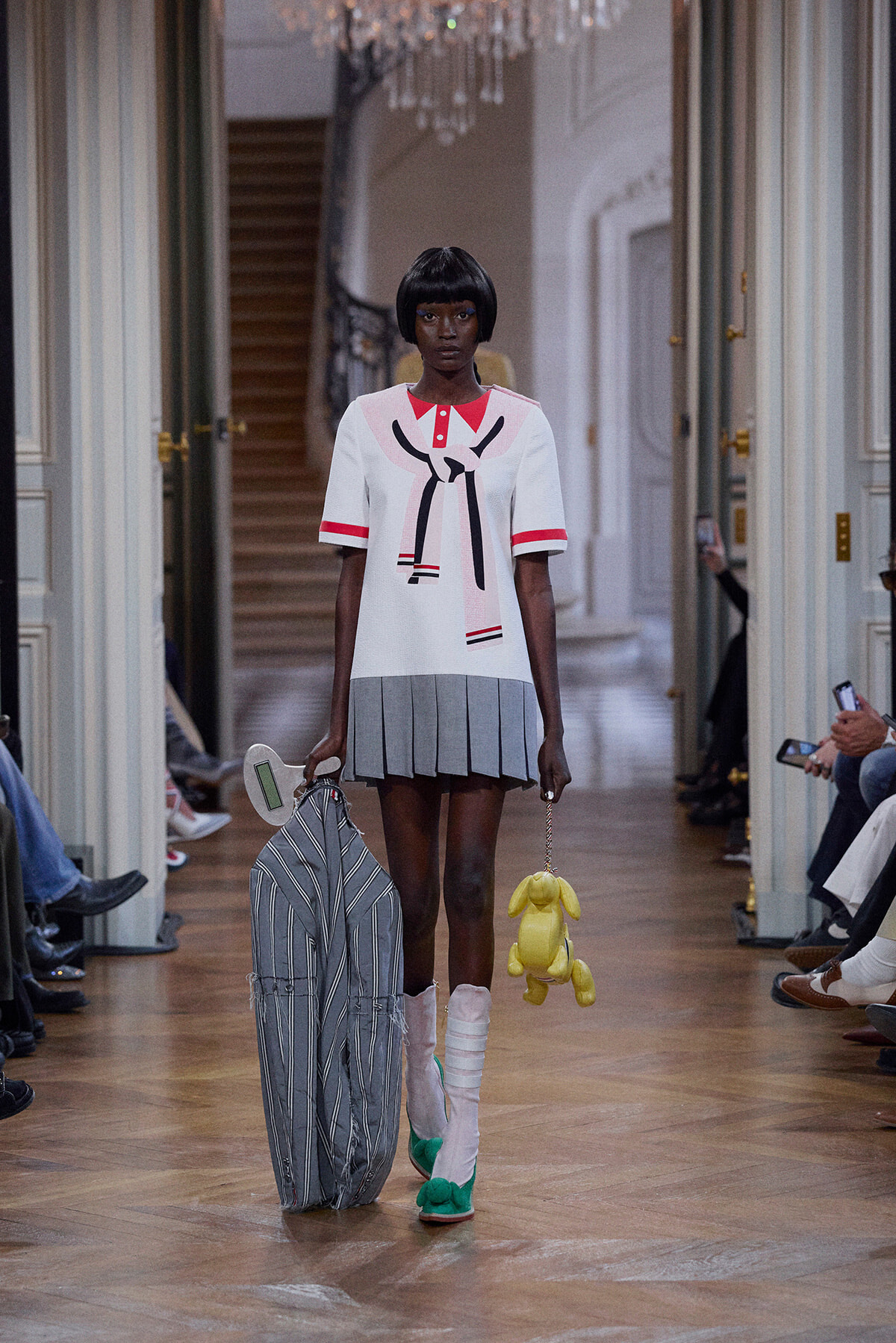 Model on a runway wearing a white top with a bow design and a pleated gray mini-skirt, carrying a large striped bag and a yellow plush bunny.