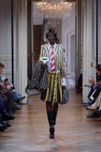 Model walks a runway in a yellow-blue striped suit with a red striped tie, carrying a large striped bag in a grand hallway. chandelier above.