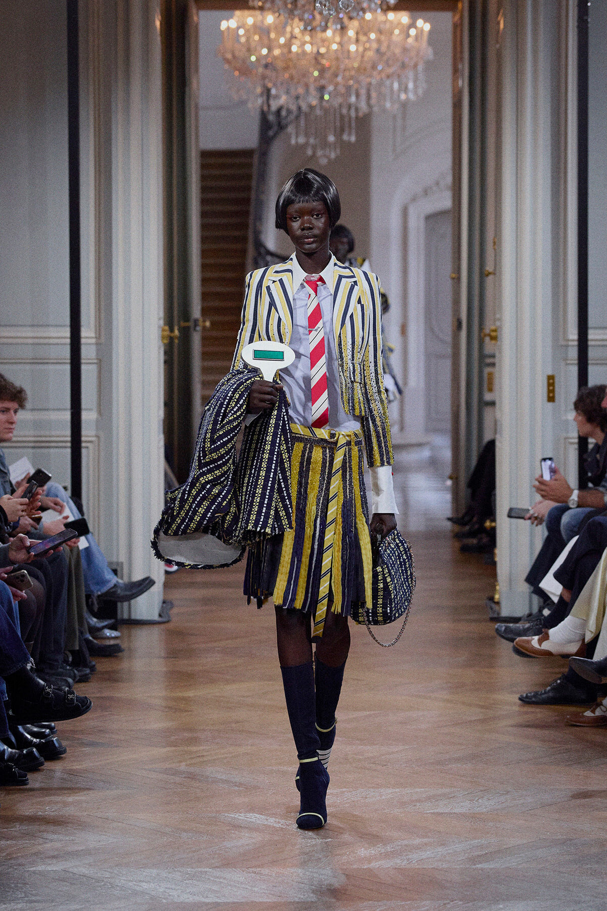 Model walks a runway in a yellow-blue striped suit with a red striped tie, carrying a large striped bag in a grand hallway. chandelier above.