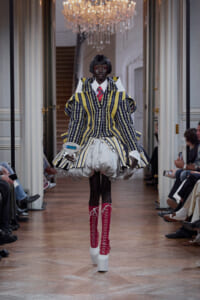 Model walks a runway in a bold striped suit with oversized shoulders and a voluminous white skirt, pink knee-high lace-up boots, chandeliers above.