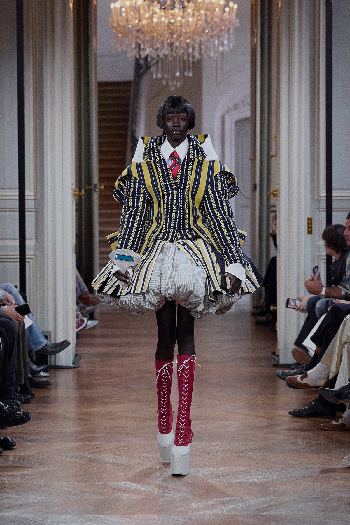 Model walks a runway in a bold striped suit with oversized shoulders and a voluminous white skirt, pink knee-high lace-up boots, chandeliers above.