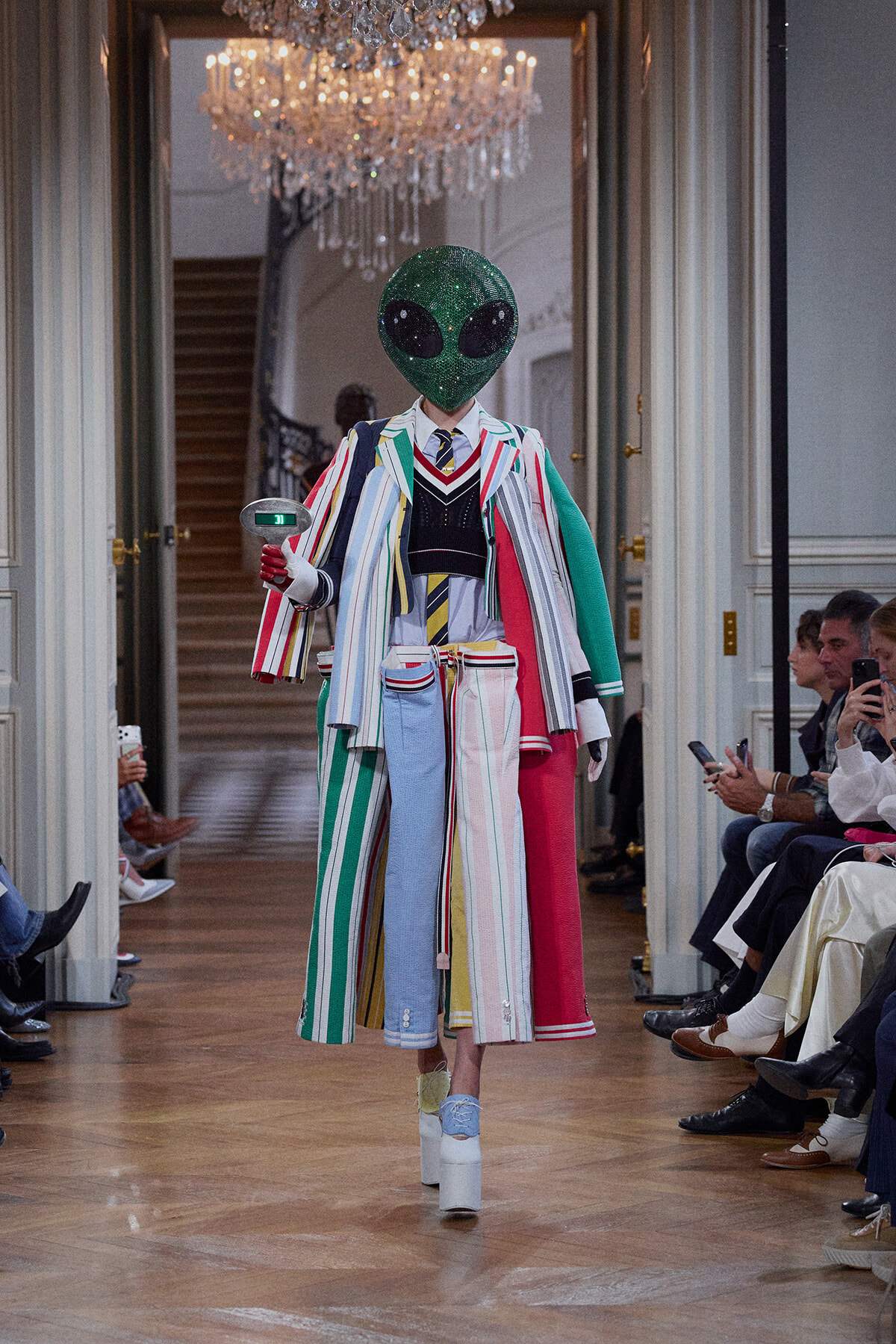 Model on a runway in a bright, multicolored striped suit and glittery green alien mask, walking past seated audience in a chandelier-lit hallway.