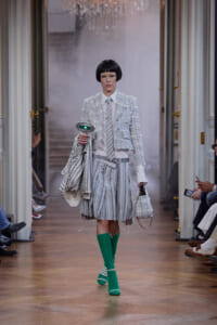 Model walks a fashion runway in a light gray plaid cropped jacket, white shirt, striped tie, and a voluminous striped skirt with bright green knee-high socks and matching shoes, carrying a striped bag and small tote.