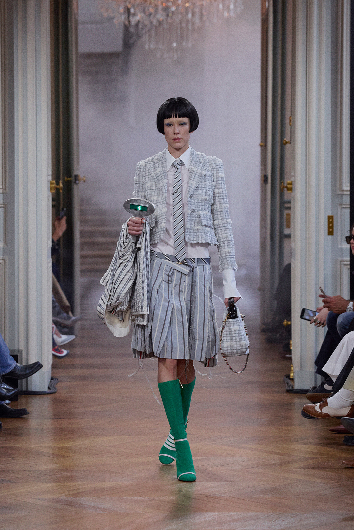 Model walks a fashion runway in a light gray plaid cropped jacket, white shirt, striped tie, and a voluminous striped skirt with bright green knee-high socks and matching shoes, carrying a striped bag and small tote.