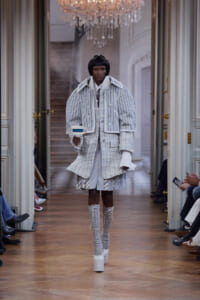 Runway model walks a fashion show in a white and gray layered ensemble with a striped cropped jacket and tall lace-up boots, holding a small rectangular accessory centerpiece.