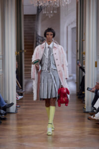 Model walks the runway in a pale pink coat over a gray striped outfit, yellow knee-high socks, and yellow-heeled shoes, carrying a red plush dog and a handheld mirror in a grand hallway setting.