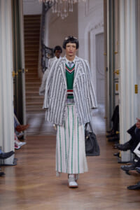 Model walks a runway in a loose gray-and-white striped suit over a green sweater and wide striped skirt, carrying a gray bag.