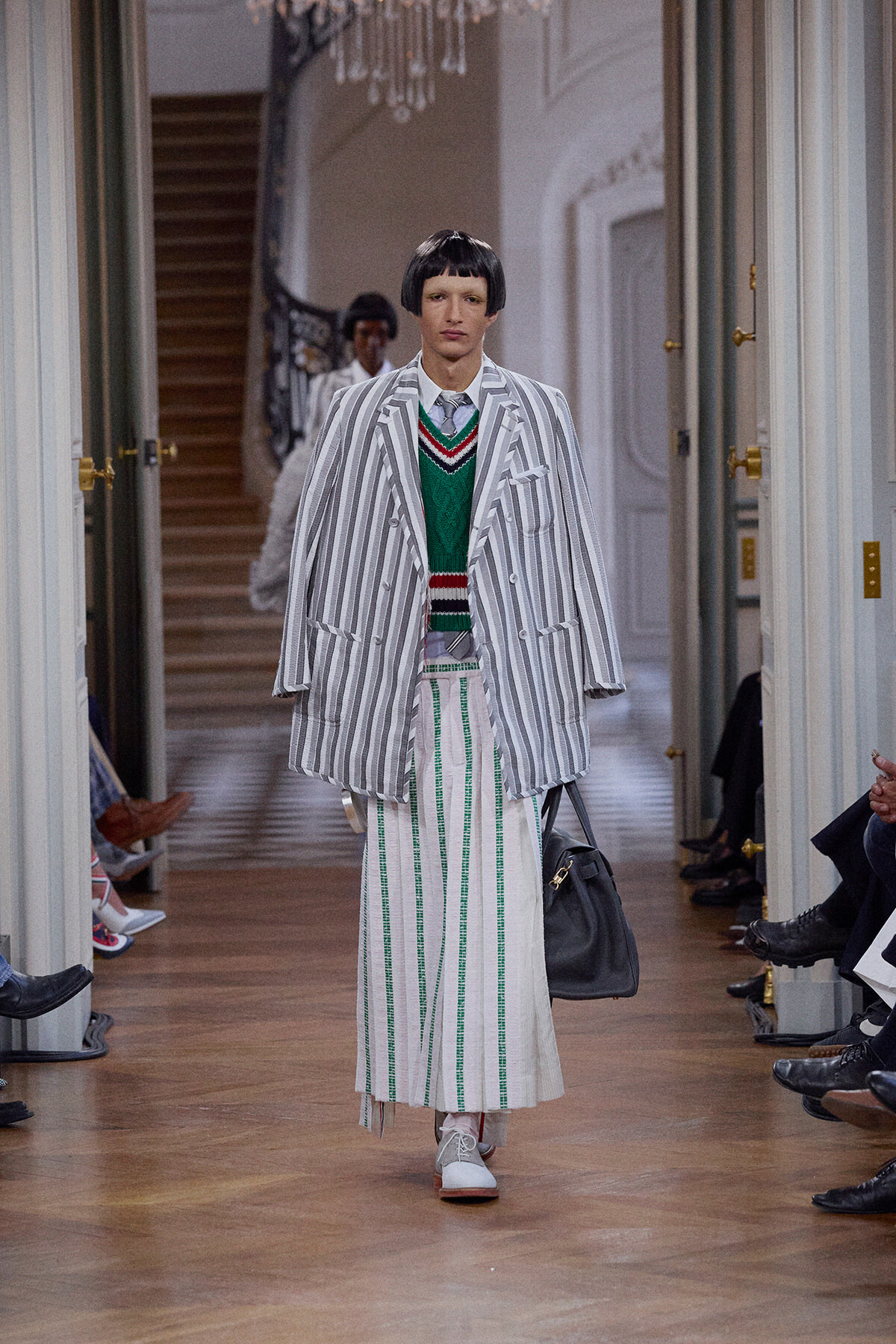 Model walks a runway in a loose gray-and-white striped suit over a green sweater and wide striped skirt, carrying a gray bag.