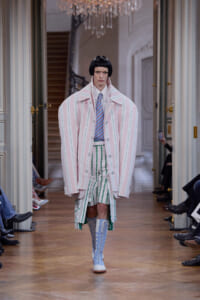 Runway model wearing an oversized pale pink striped coat over a striped shirt and green-striped shorts, with blue lace-up boots.