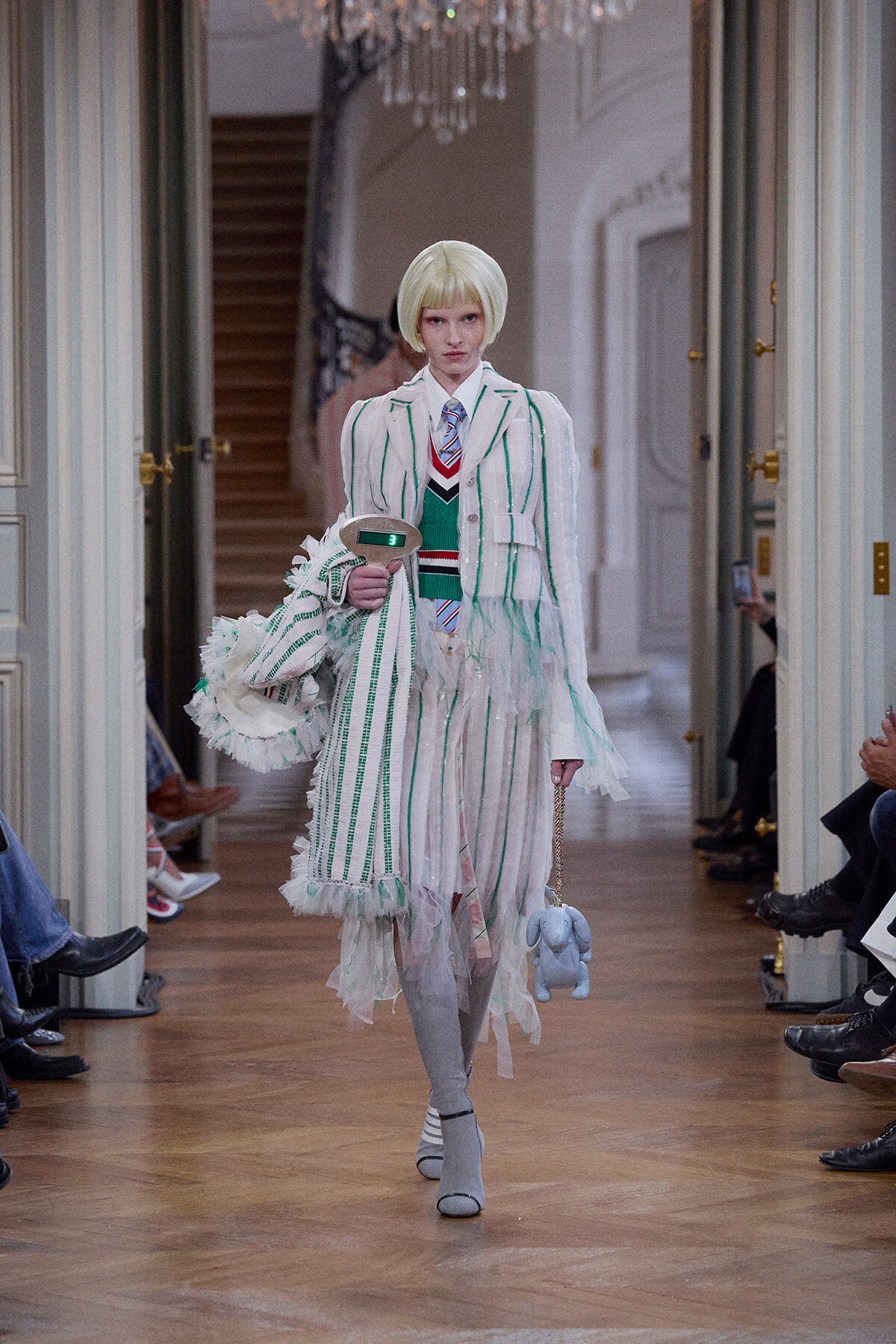 Model walks a runway in a white and green striped fringe suit, holding a matching coat and a blue elephant plush on a chain.