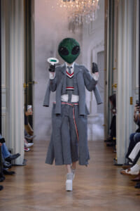 Model in a gray layered suit walking a runway, wearing a sparkly green alien mask and white platform heels, holding a small prop device.