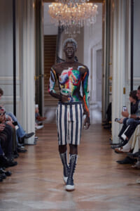 Black male model walks a fashion runway in a multicolored sequined long-sleeve top, striped knee-length shorts, and tall lace-up boots, in an ornate venue with a chandelier.