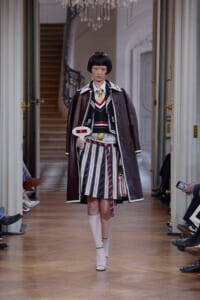 Model on a runway wearing a layered school-uniform-inspired outfit: striped pleated skirt, vest, white shirt, cardigan, and a long dark coat.