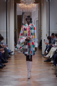 Model walking a fashion runway in a bright, patchwork suit with a matching bag and white knee-high boots.