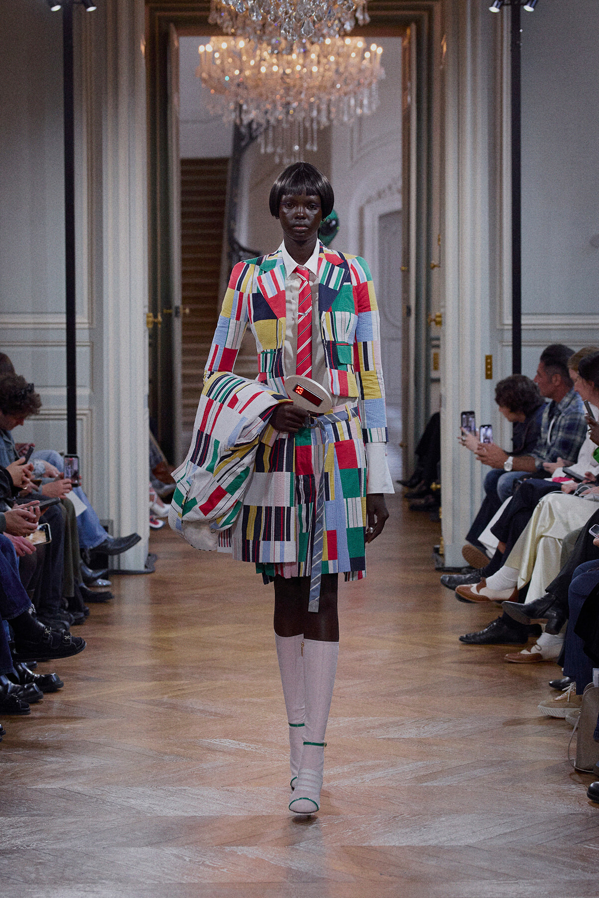 Model walking a fashion runway in a bright, patchwork suit with a matching bag and white knee-high boots.