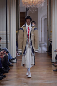Model walks runway in oversized beige coat with navy trim over white shirt and red tie, layered skirt.