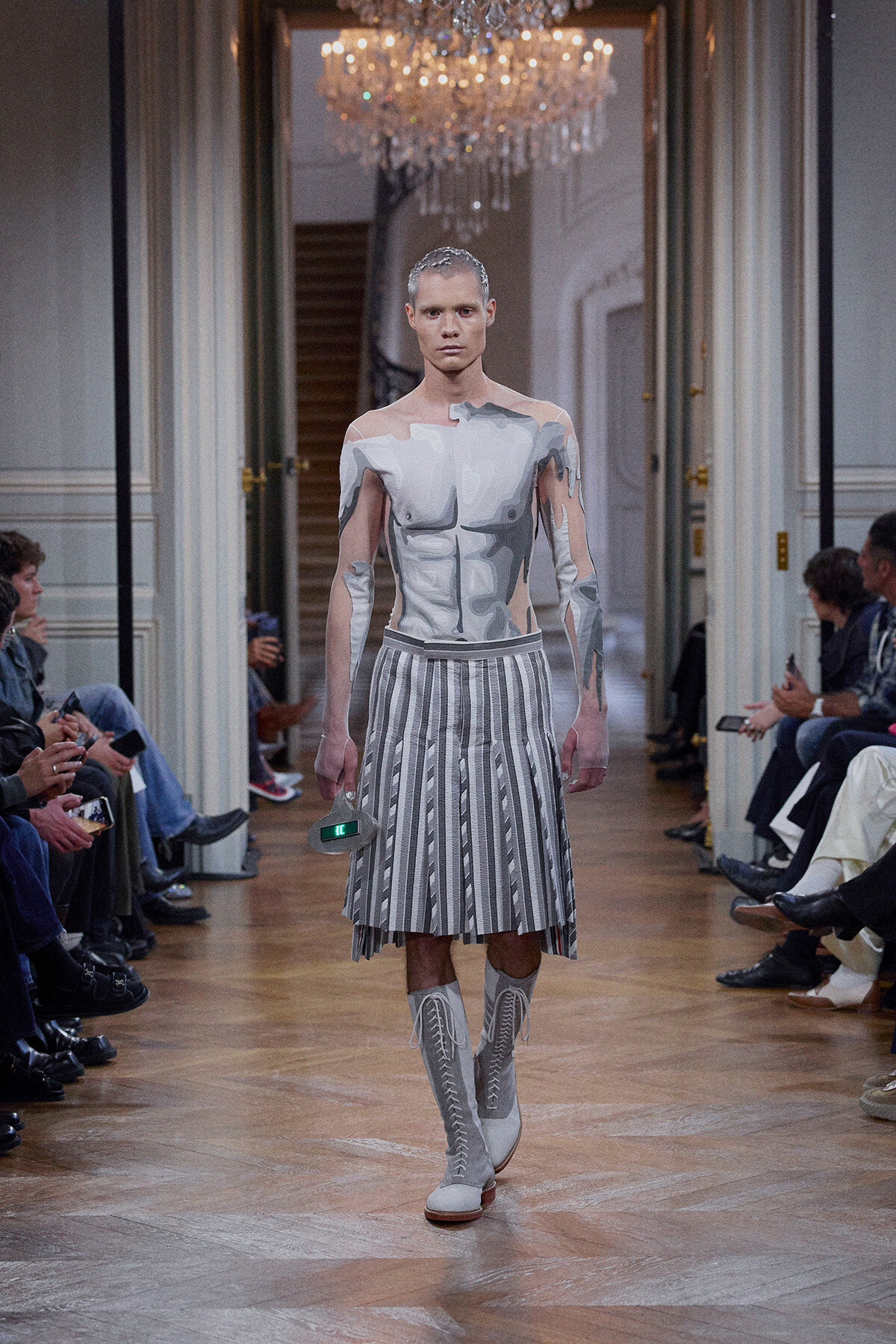 Model walks the runway in a gray armor-printed bodysuit, a striped pleated skirt, and knee-high lace-up boots.