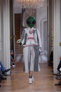 Model walks a runway in a two-tone gray and white suit with pink knit vest, wearing a glittery green alien head mask and white platform shoes under chandeliers.
