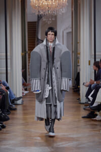 Model walks a fashion runway in a voluminous, layered gray coat with striped cuffs and tall collar.