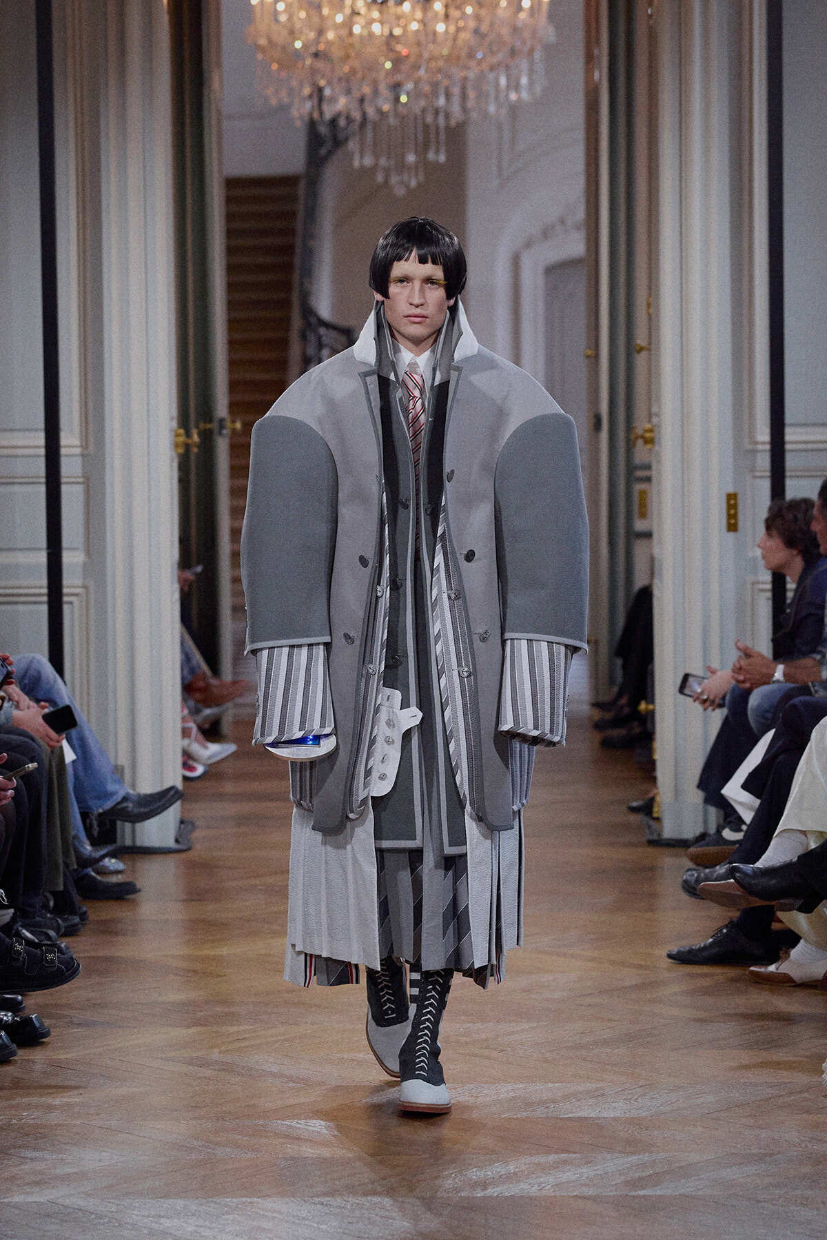 Model walks a fashion runway in a voluminous, layered gray coat with striped cuffs and tall collar.