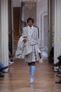 Model on a runway wearing a gray-and-white striped suit, carrying a textured gray coat and a pale pink handbag in a grand hallway.