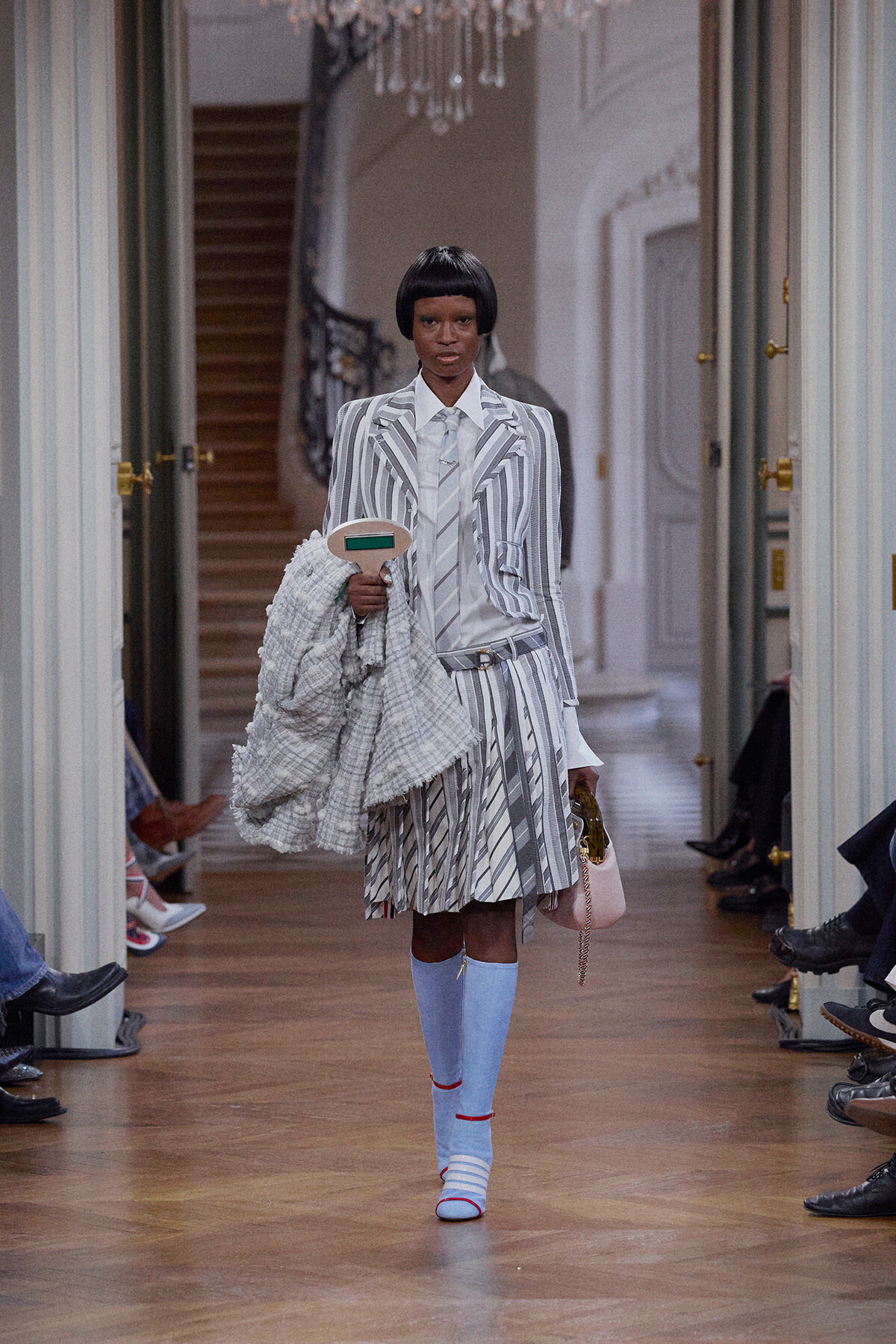 Model on a runway wearing a gray-and-white striped suit, carrying a textured gray coat and a pale pink handbag in a grand hallway.