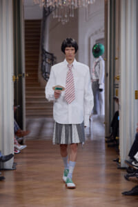 Model walks a runway in a white oversized shirt, striped tie, and pleated gray skirt, wearing mint-green sneakers and gray socks, holding a small handheld device in a hallway with a chandelier in the background
