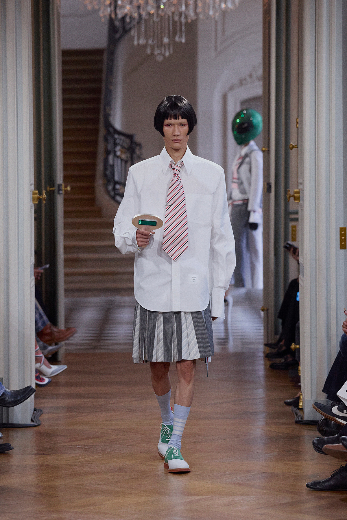 Model walks a runway in a white oversized shirt, striped tie, and pleated gray skirt, wearing mint-green sneakers and gray socks, holding a small handheld device in a hallway with a chandelier in the background