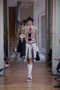 Model walks a runway in a beige blazer, striped sweater, and white pleated skirt with knee-high socks, carrying a navy coat and black handbag.