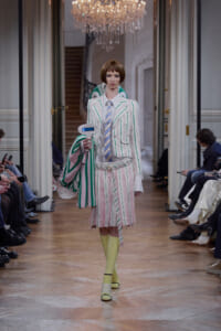 Model walks a runway in a white pinstripe suit with green and pink stripes, carrying a large striped tote and a blue handheld device.
