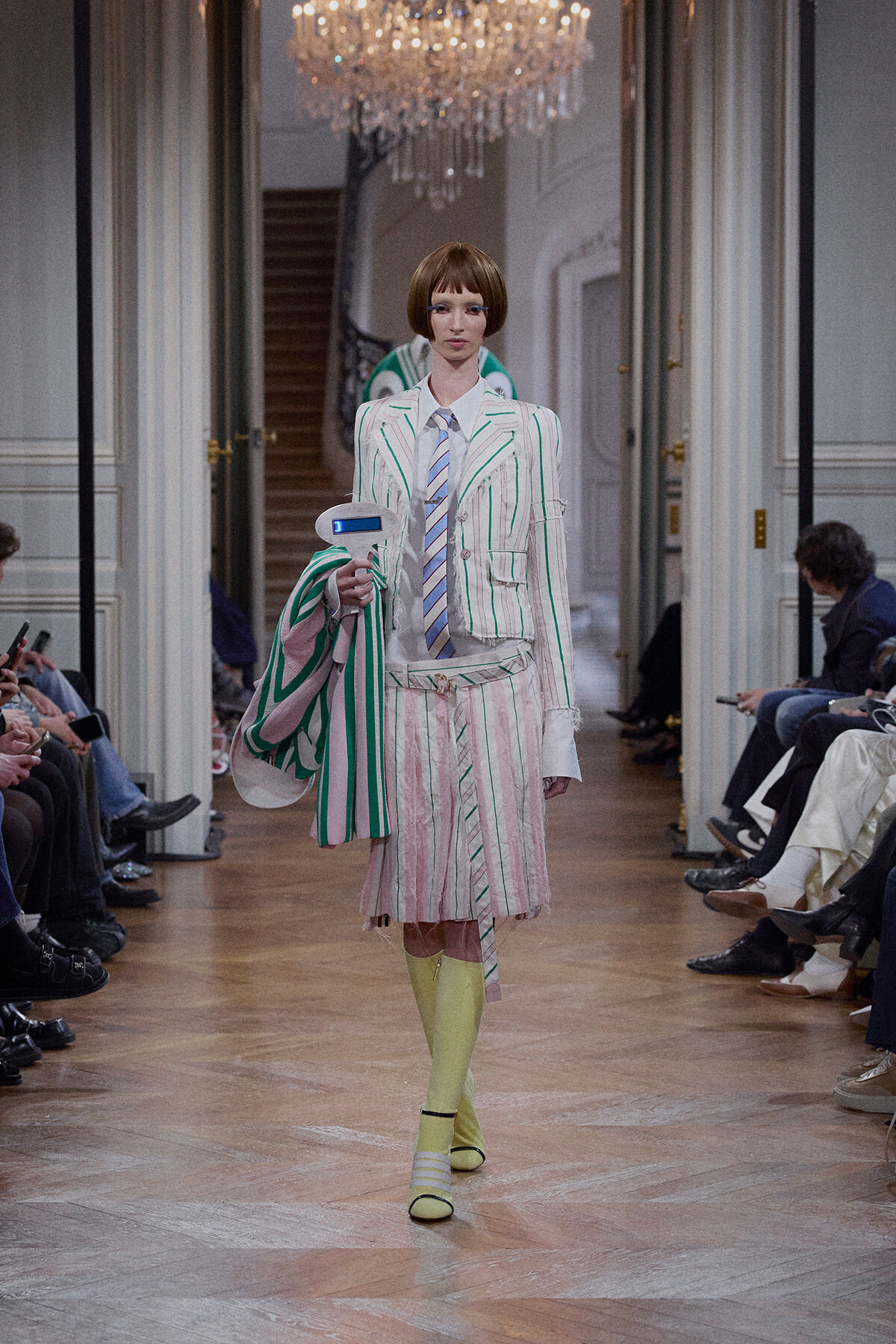 Model walks a runway in a white pinstripe suit with green and pink stripes, carrying a large striped tote and a blue handheld device.