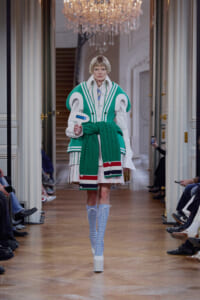 Model walks a runway in a bold green and white oversized vest with a chunky knit green belt, holding a blue card; chandeliers and a grand hallway backdrop.
