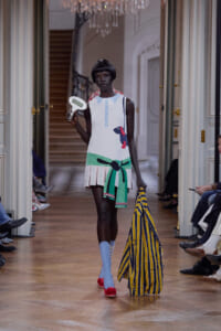 Runway model wearing a white sleeveless dress with a dog graphic, green sash and fringe, blue socks, red shoes, carrying a yellow‑blue striped bag and a handheld scale.