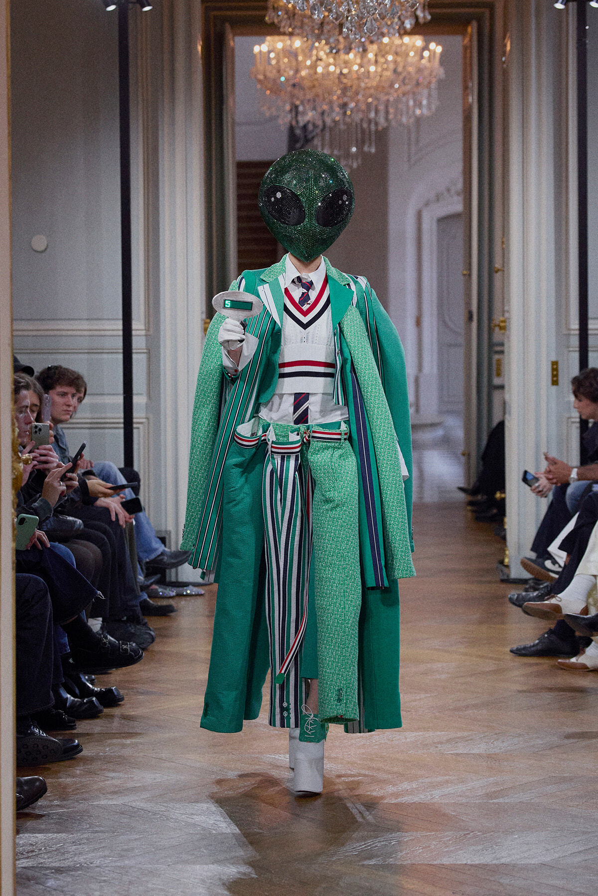 Model walks a fashion runway in a green, multi-patterned suit with a glittery alien head mask, audience seated along the catwalk.